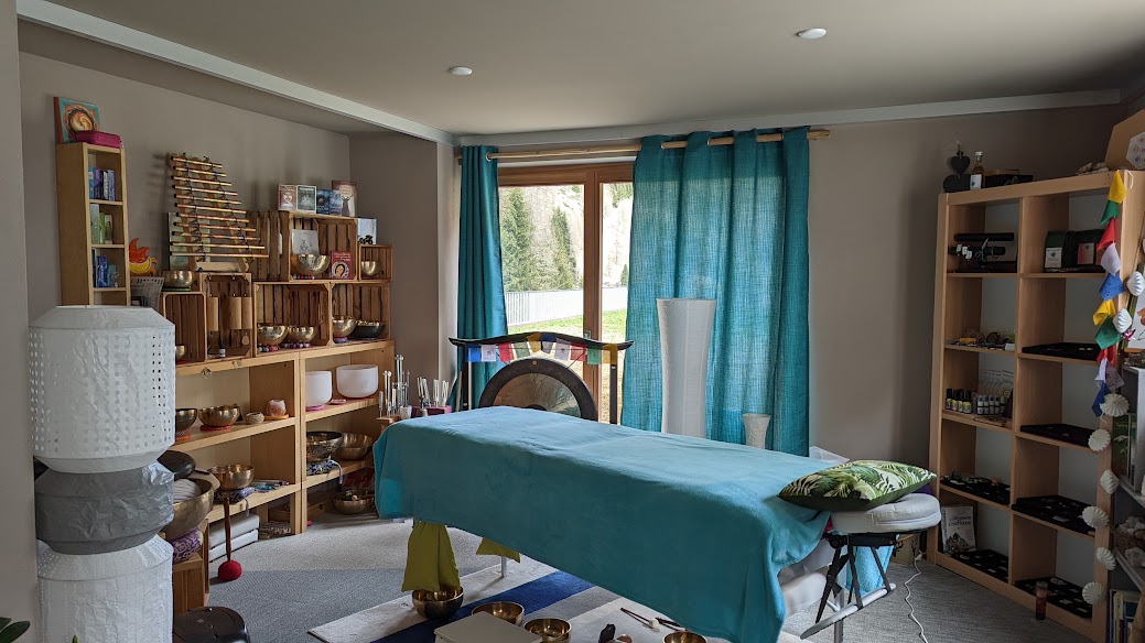 Intérieur d'un espace de bien-être avec une table de massage au centre, des étagères chargées d'objets de relaxation et des rideaux turquoise laissant entrer la lumière naturelle.